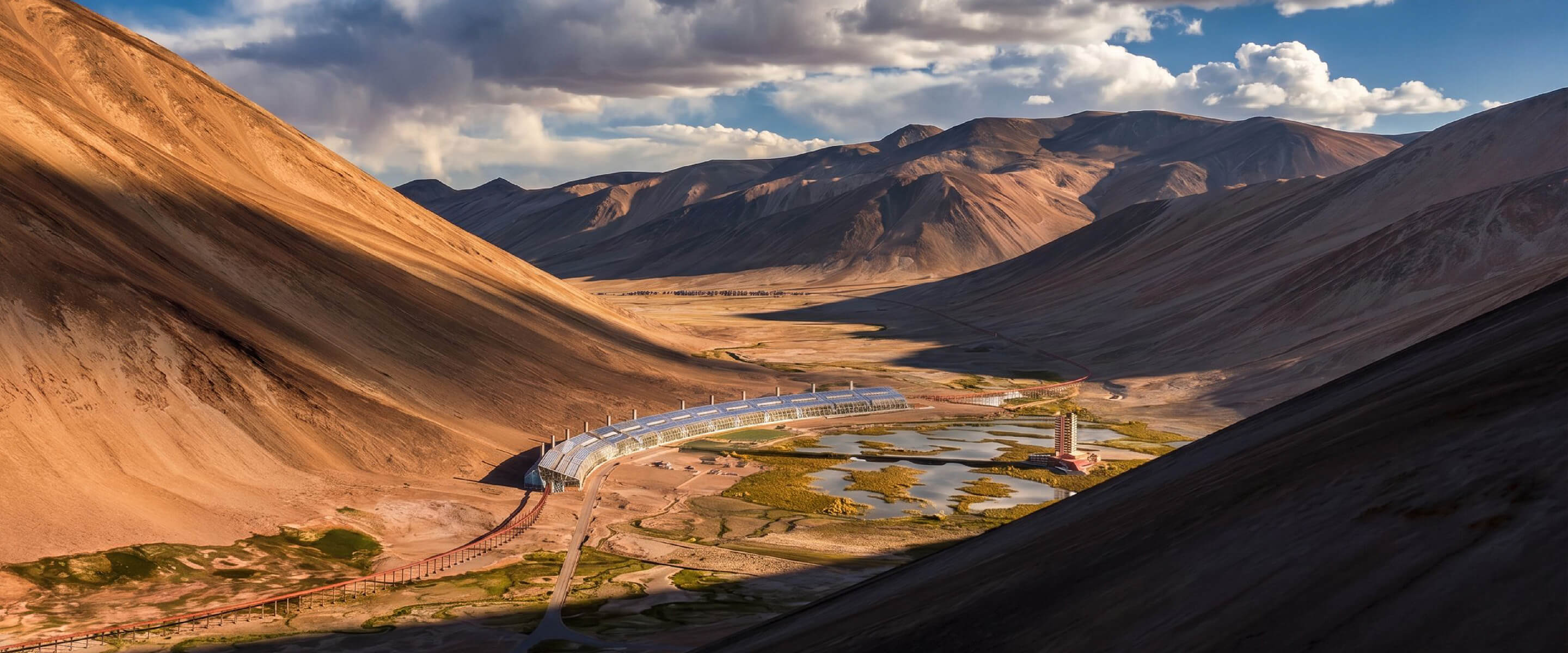 Aerial view of a curved building nestled between mountains, surrounded by a landscape of valleys and water bodies under a cloudy sky.