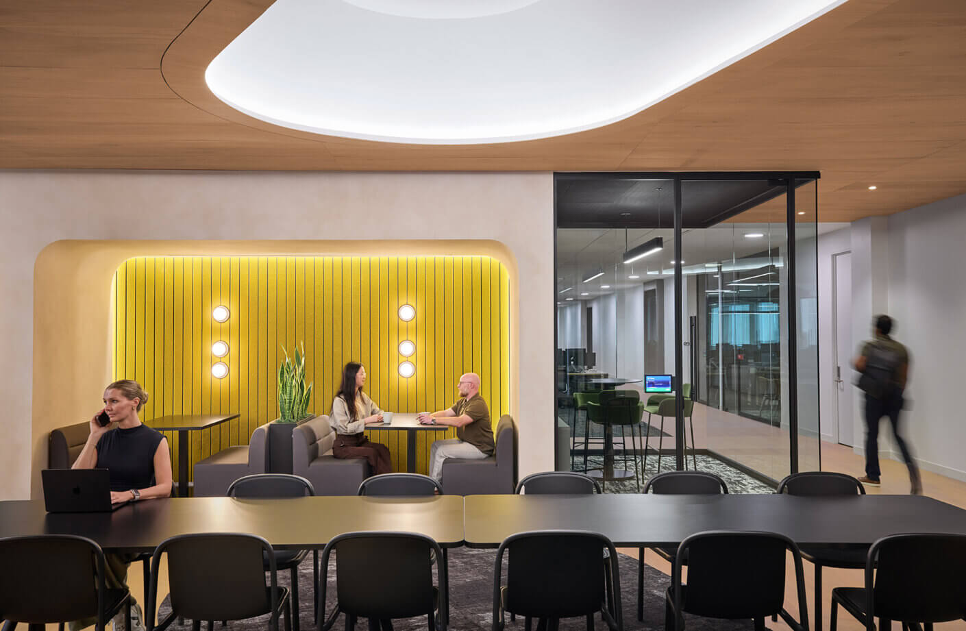 Interior office space featuring a yellow accent wall, seating areas, and glass-walled meeting rooms with people engaged in conversations and work.