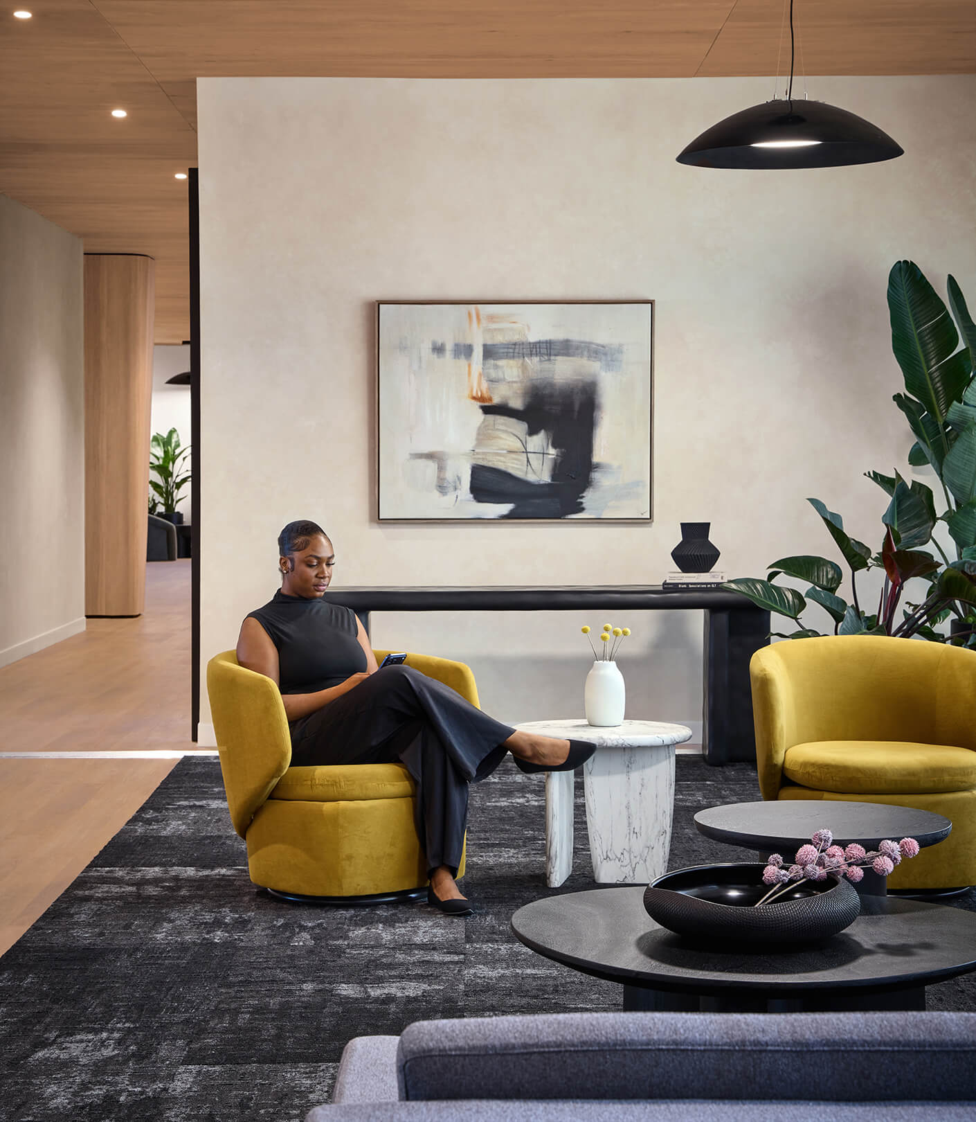 A woman sits in a yellow chair, focused on her phone, surrounded by contemporary furniture and indoor plants in a well-lit space.