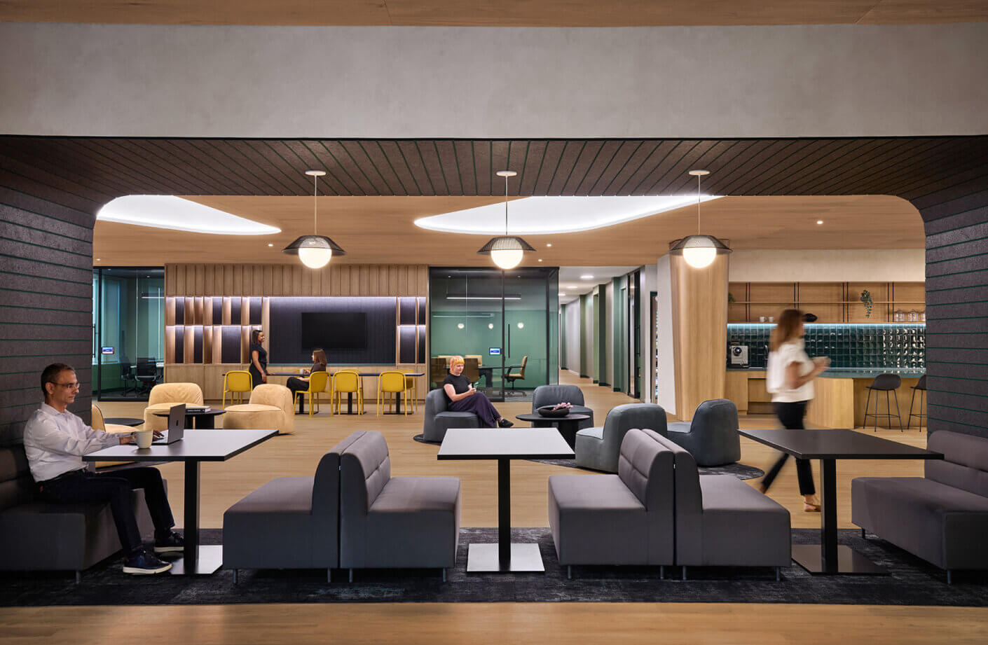 Office interior featuring seating areas, a communal table, and individuals engaged in work and conversation. Natural light and stylish design elements.