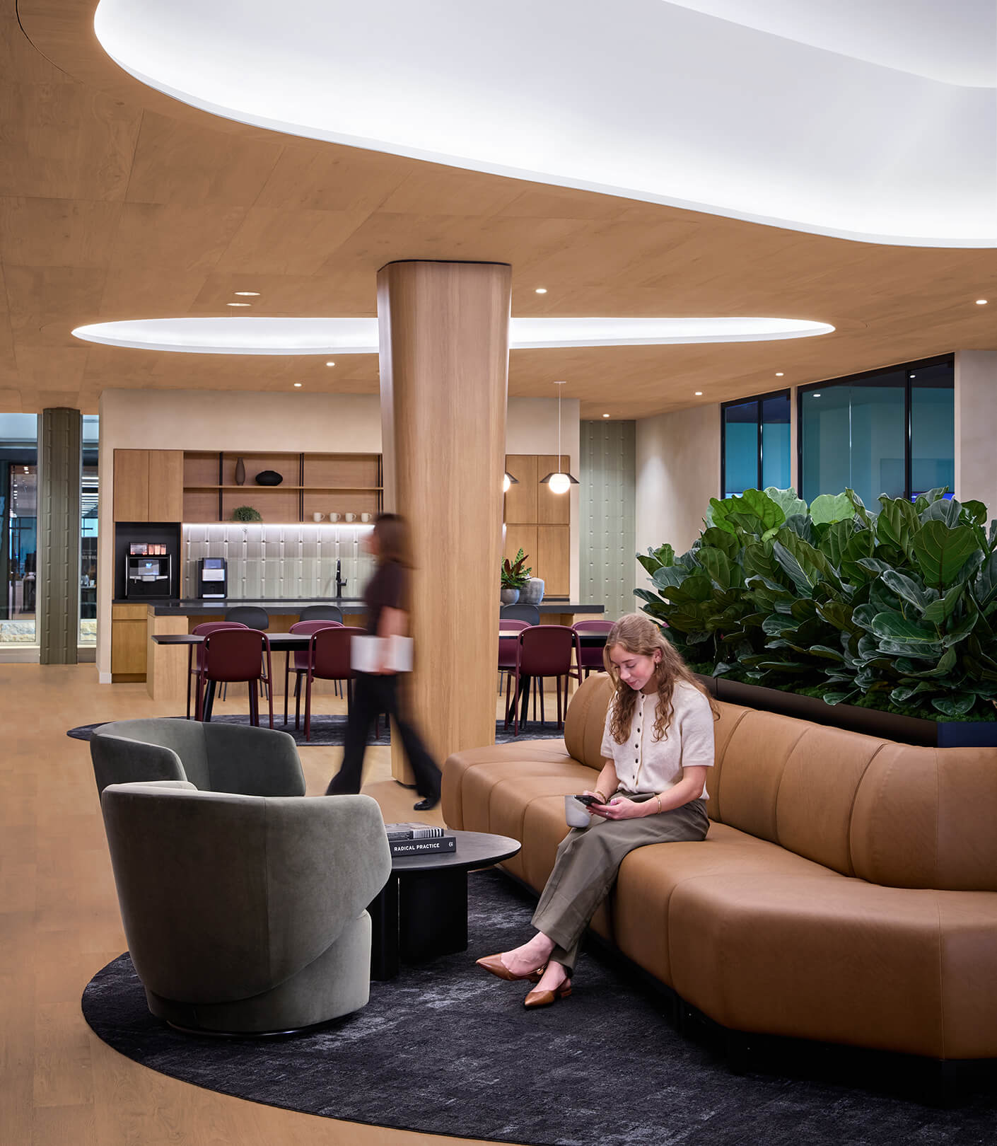 A woman sits on a leather couch using her phone, with modern furniture and lush plants in a well-lit interior space.