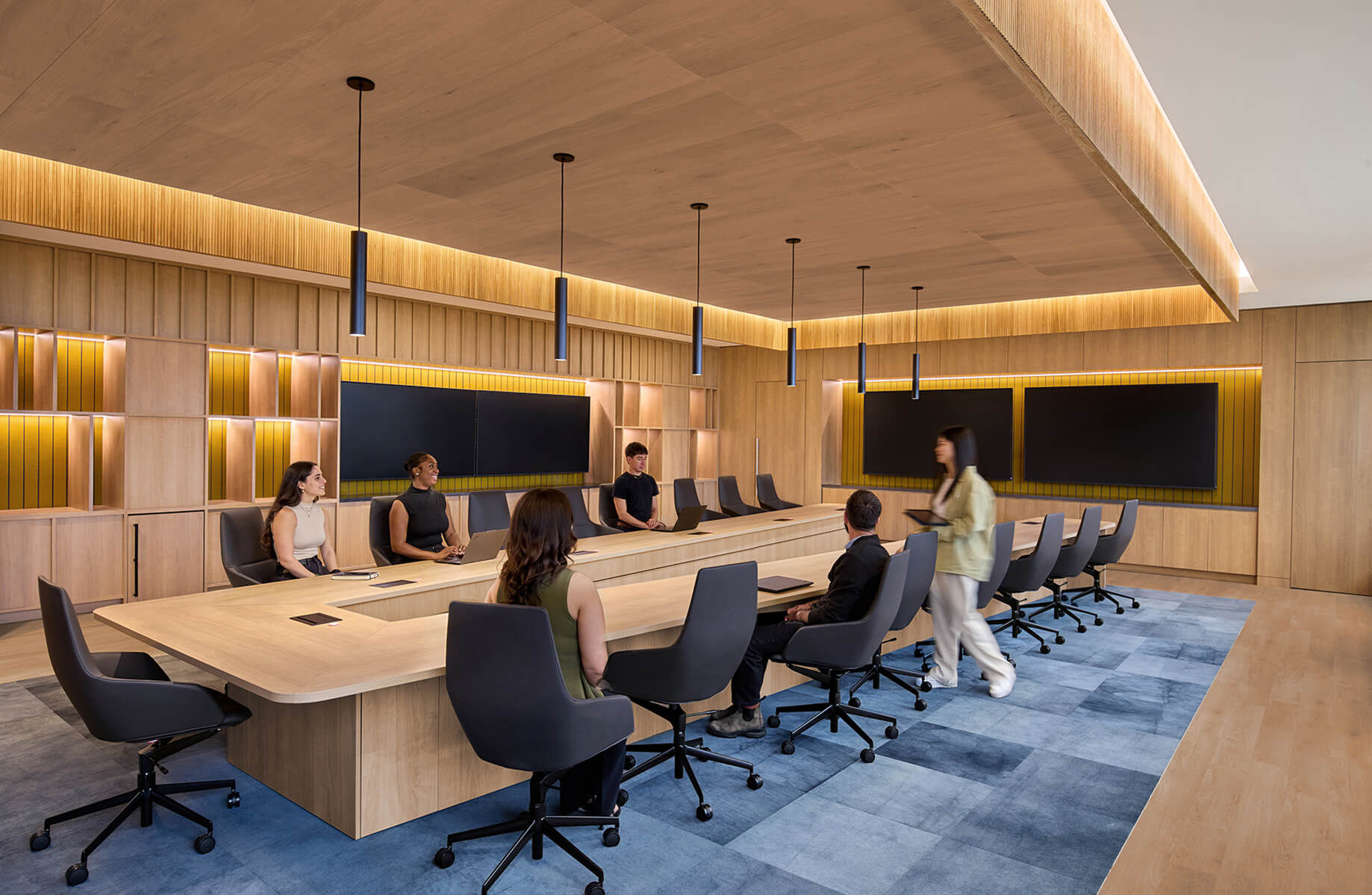 A spacious conference room featuring a long wooden table, modern seating, and multiple screens, with eight people engaged in discussion.