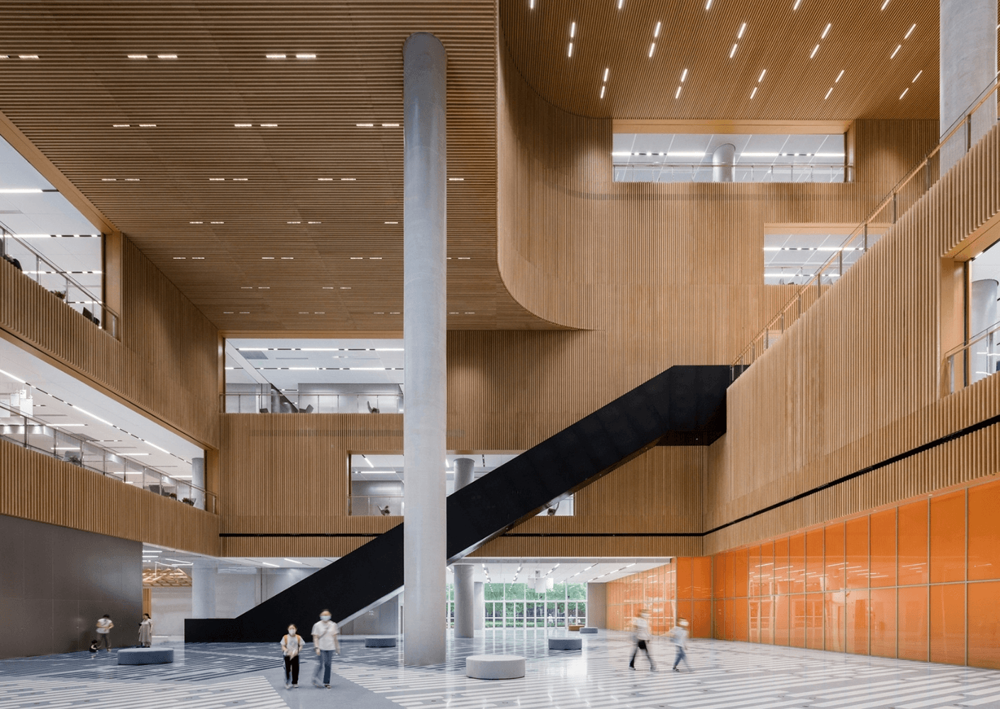 Interior view of a multi-level building with wooden accents, central staircase, and visitors walking through the space. Bright and spacious atmosphere.