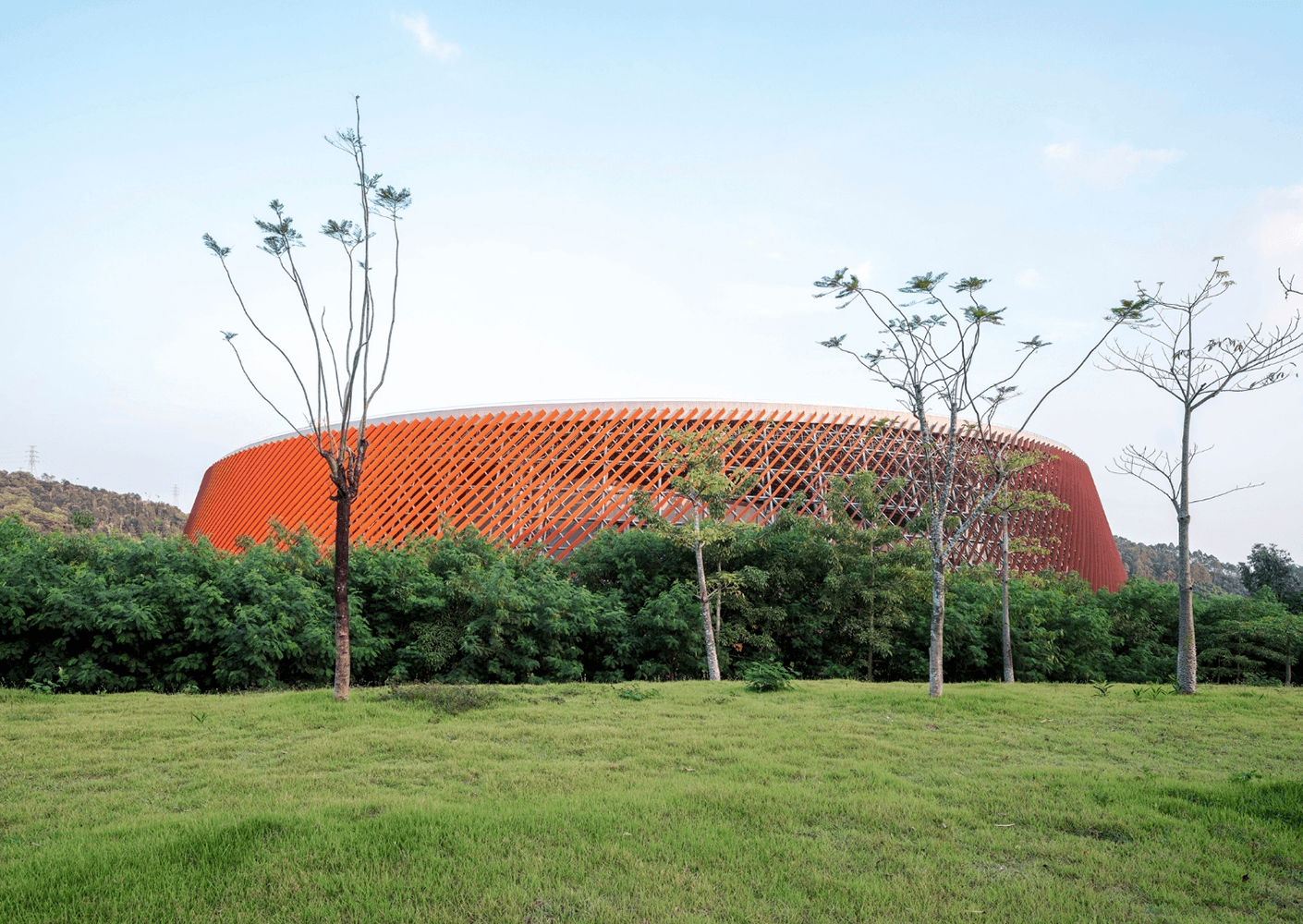 A large, circular sports arena with a distinctive orange lattice exterior, surrounded by greenery and trees against a clear sky.
