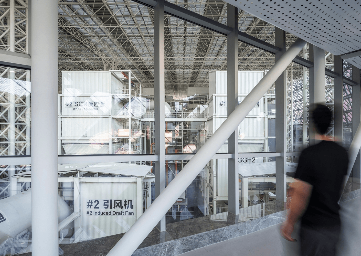 Interior view of an industrial facility showing machinery labeled with numbers. A person walks past large glass windows. Structural elements visible.