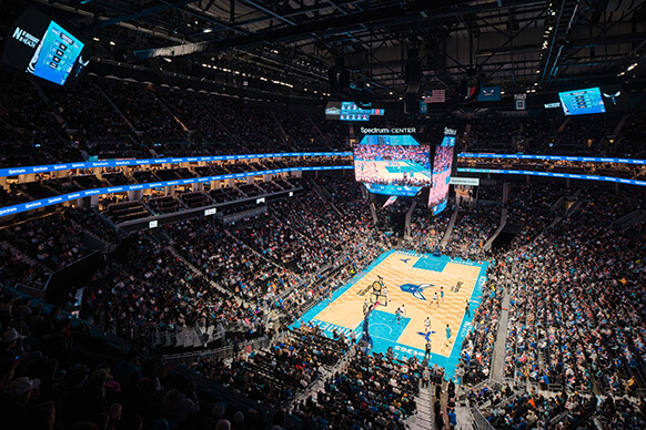 Aerial view of a packed Spectrum Center during a basketball game, featuring players on the court and a large video screen.