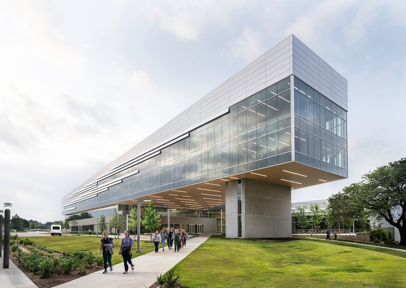 Large contemporary building with expansive glass facade, featuring a pathway and people walking in a landscaped area. Overcast sky in background.