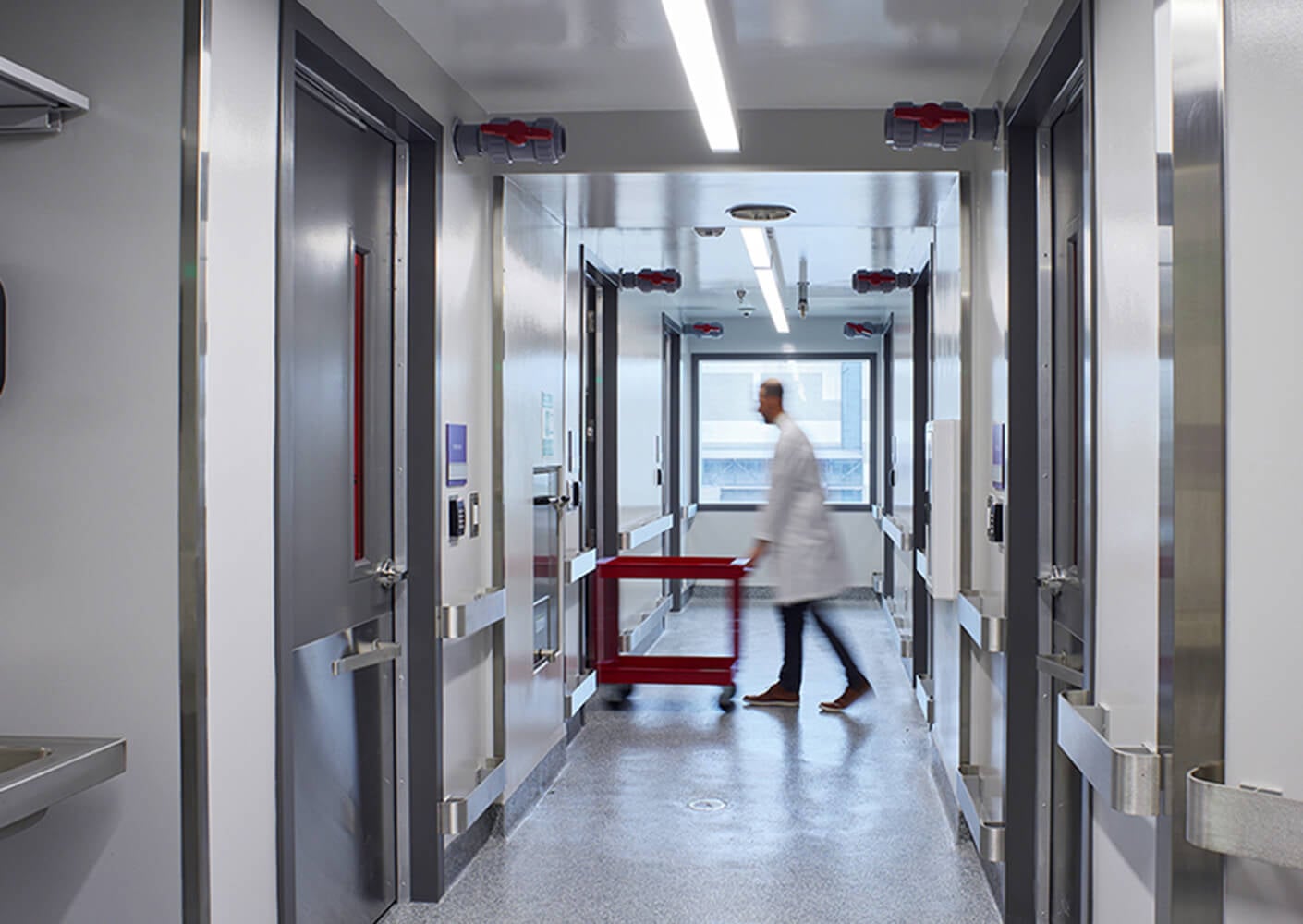 A healthcare professional walks down a corridor, pushing a red cart past doors and windows in a clinical setting.