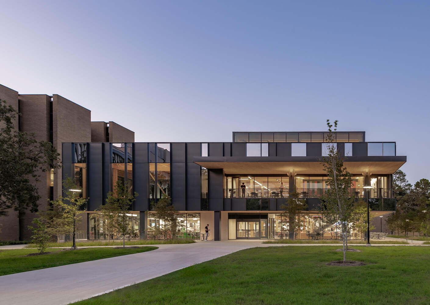 Exterior view of a building featuring large glass windows, flat roof, and surrounding landscaped area with trees and a pathway.