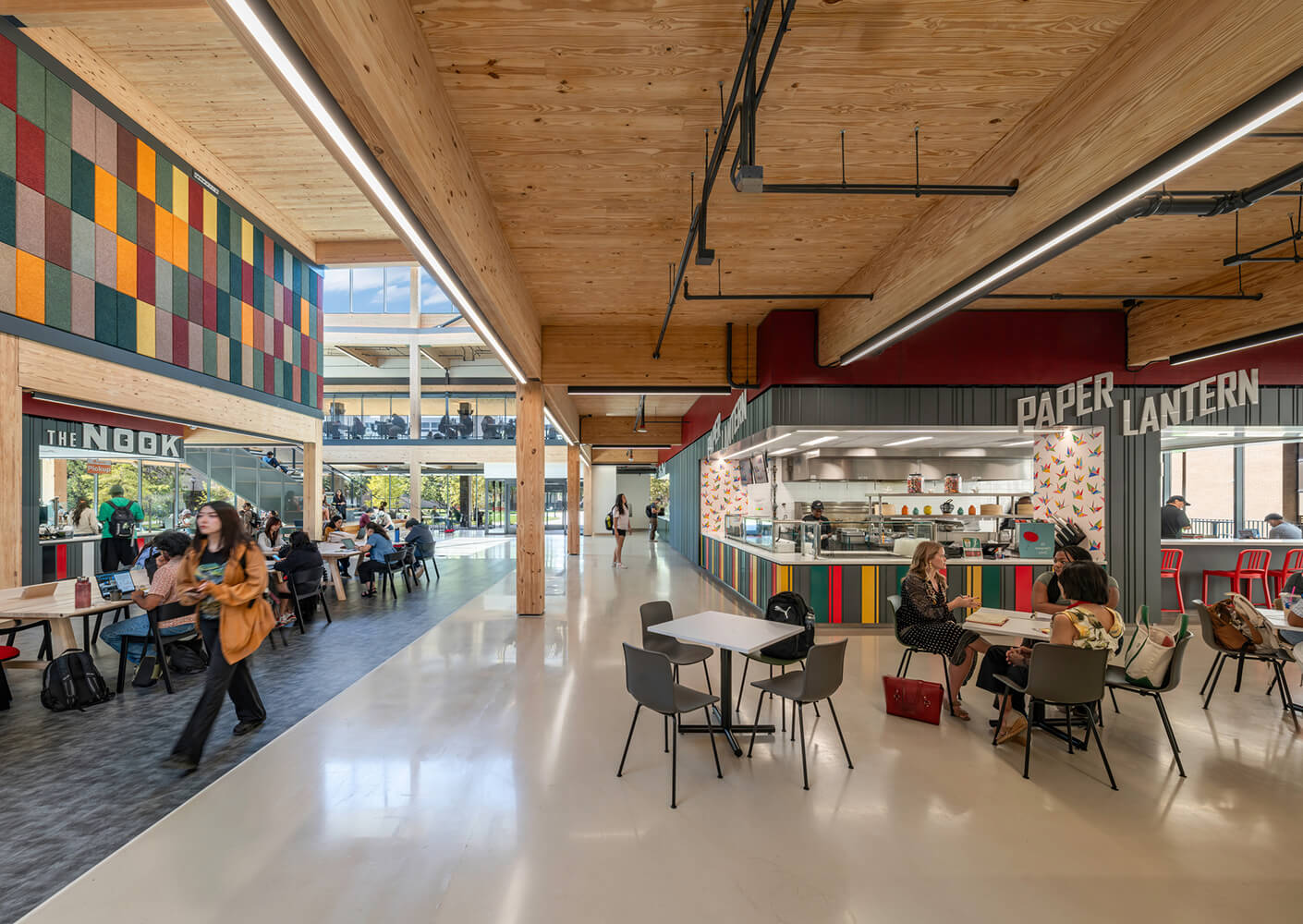 Interior view of a cafeteria with wooden beams, colorful wall panels, and dining areas where people are eating and socializing.