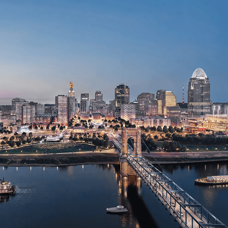 Panoramic view of Cincinnati skyline at dusk, featuring buildings, waterfront, and a bridge over the river with boats visible.