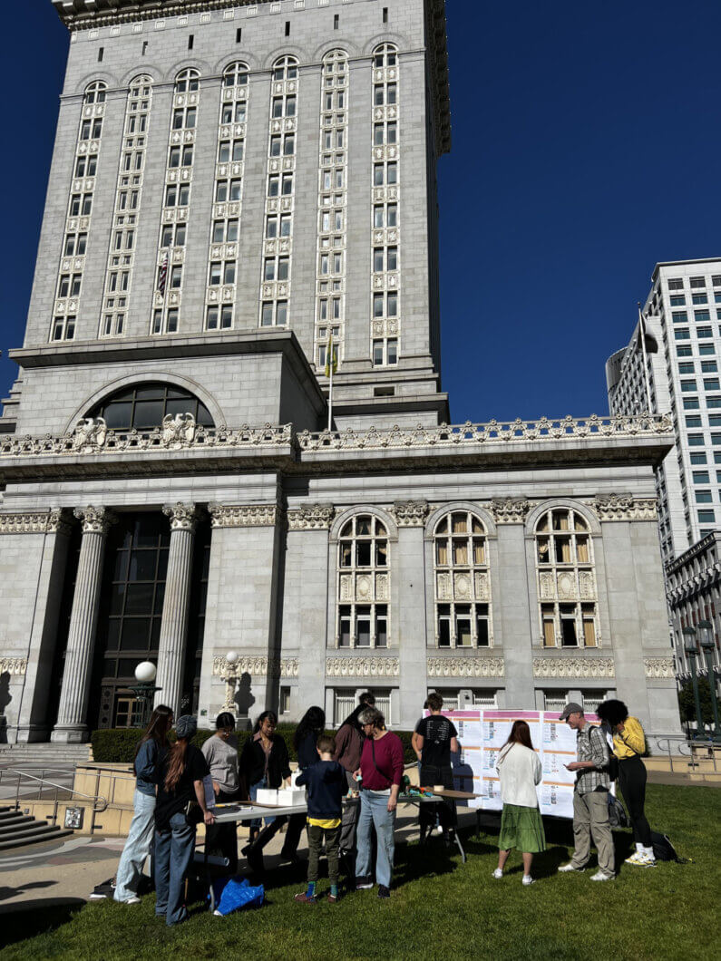 People gather around tables with displays outside a historic building under a clear blue sky. Various activities and discussions are happening.