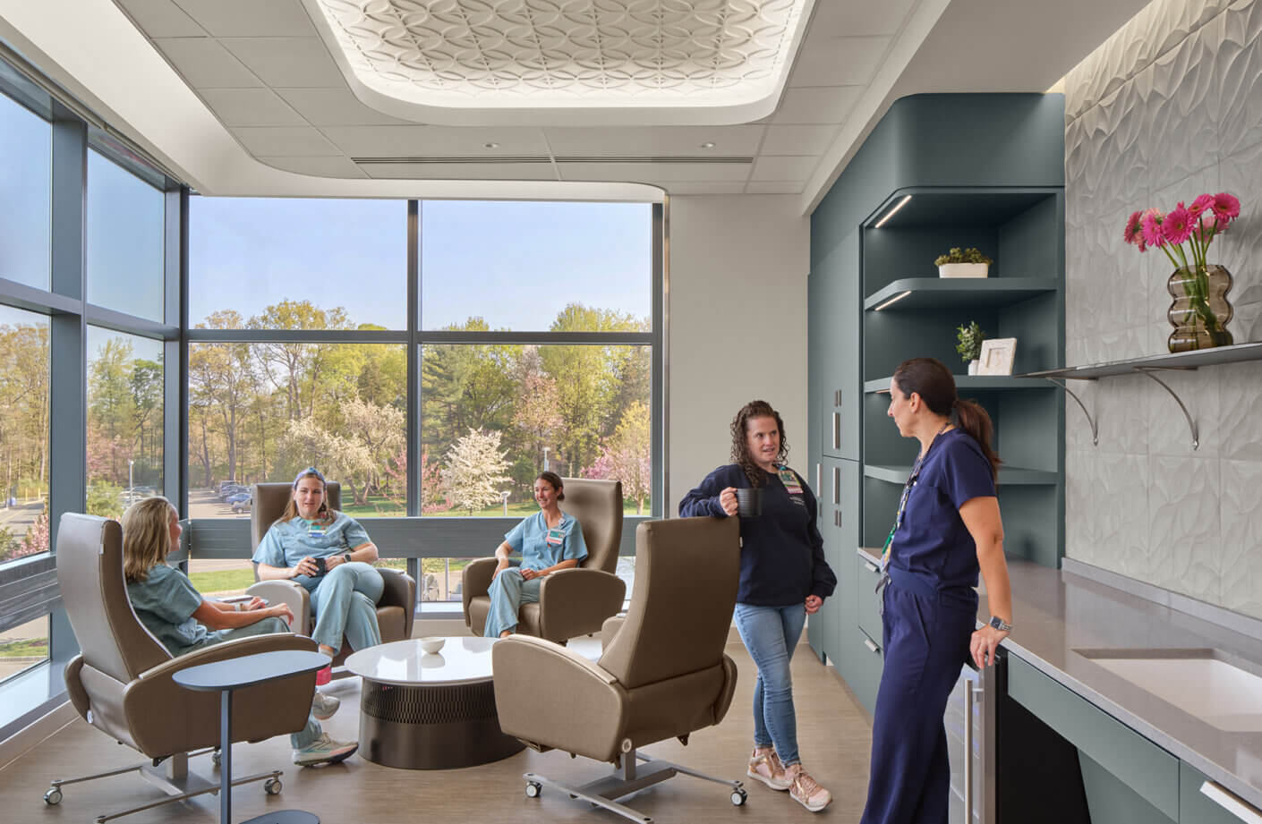 Four healthcare professionals in scrubs and casual attire engage in conversation in a well-lit lounge with large windows showcasing greenery.