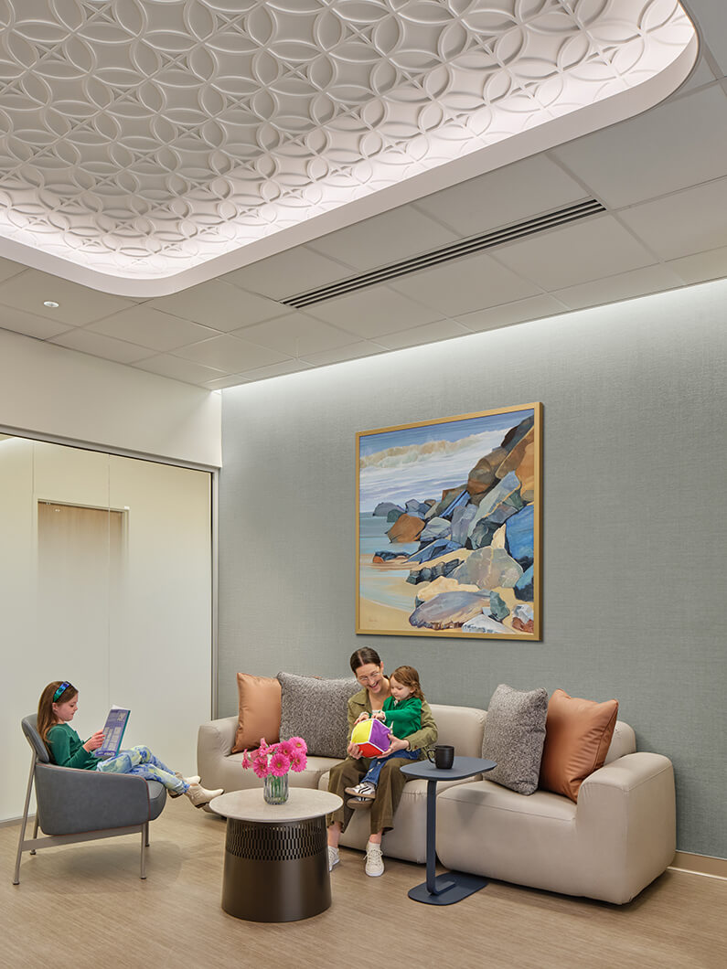 A welcoming waiting area featuring a textured ceiling, a sofa, a coffee table with flowers, and a mother with a child. A girl reads nearby.
