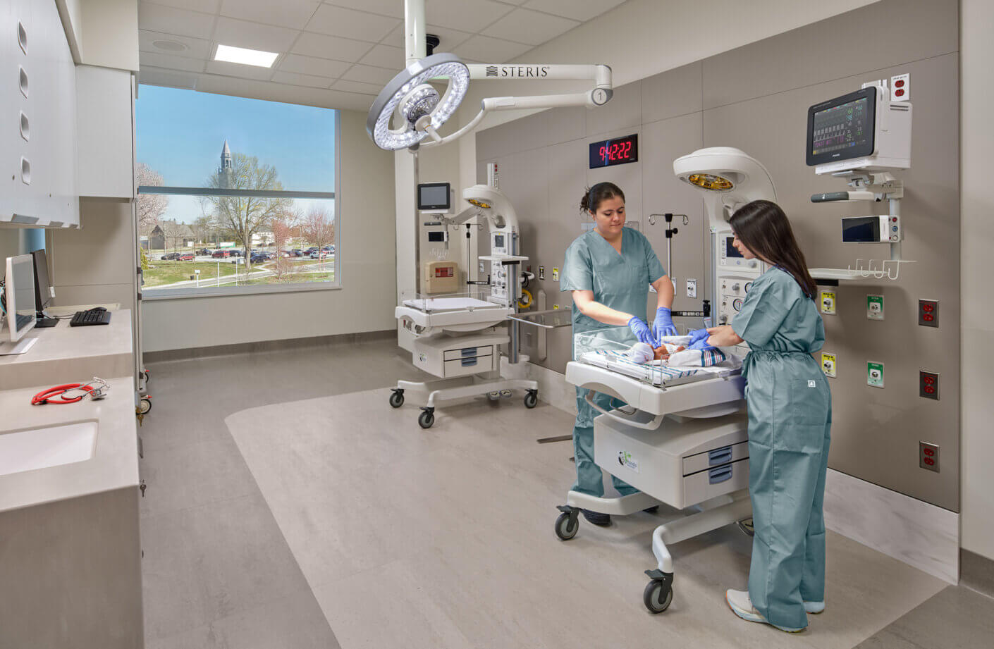 Two healthcare professionals in scrubs tending to a newborn in a neonatal care unit, equipped with medical technology and natural light from a window.
