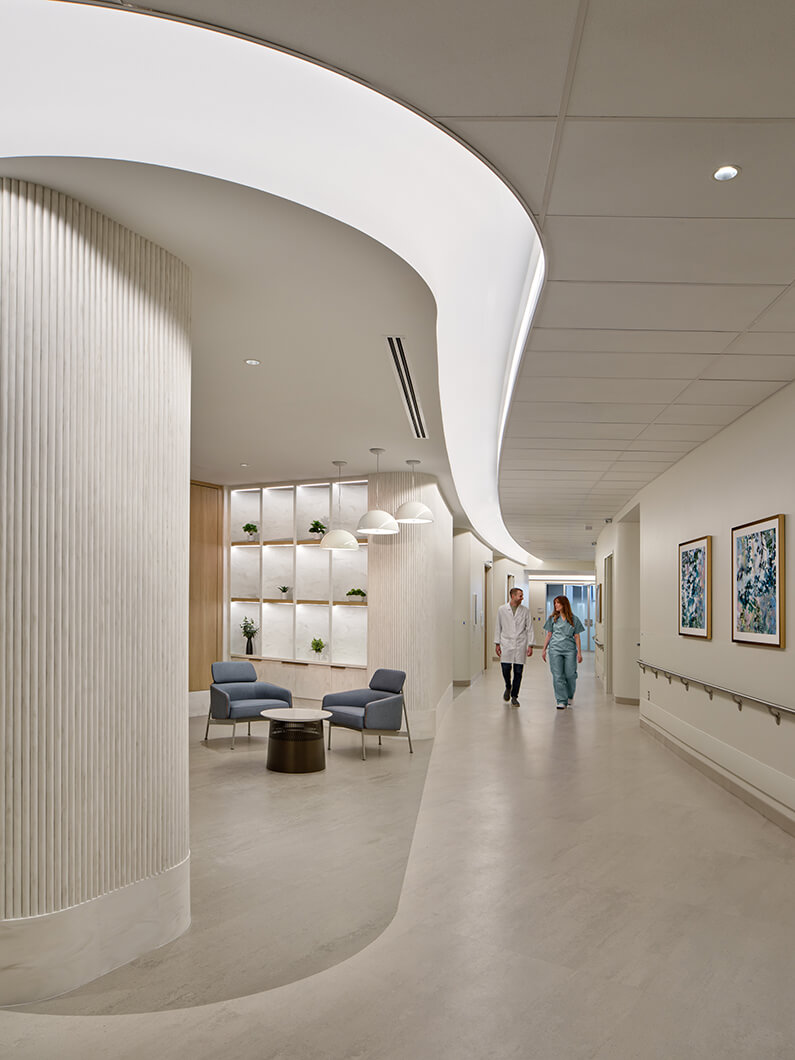 Healthcare hallway with curved ceiling, round seating area, two professionals walking, and decorative wall shelves with plants.