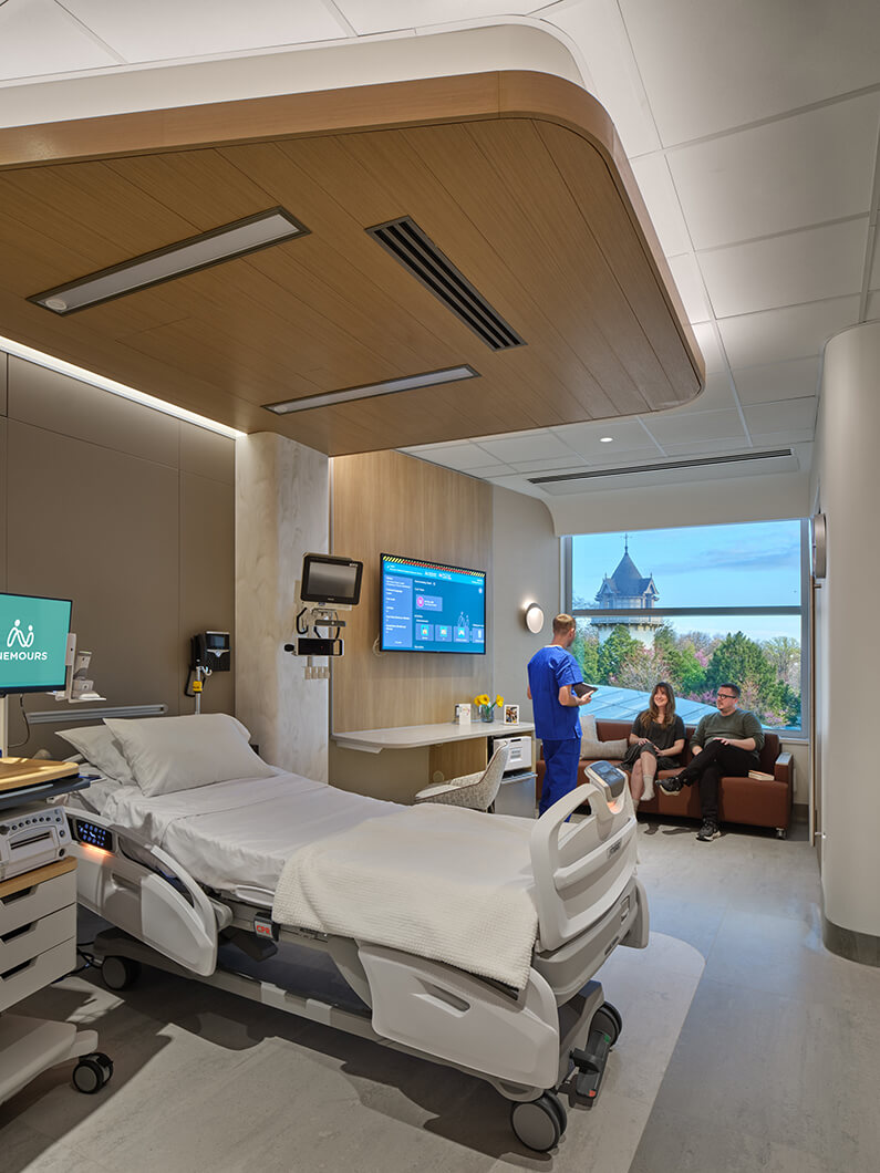 Healthcare room with a hospital bed and medical equipment. A healthcare professional interacts with two visitors near a window.
