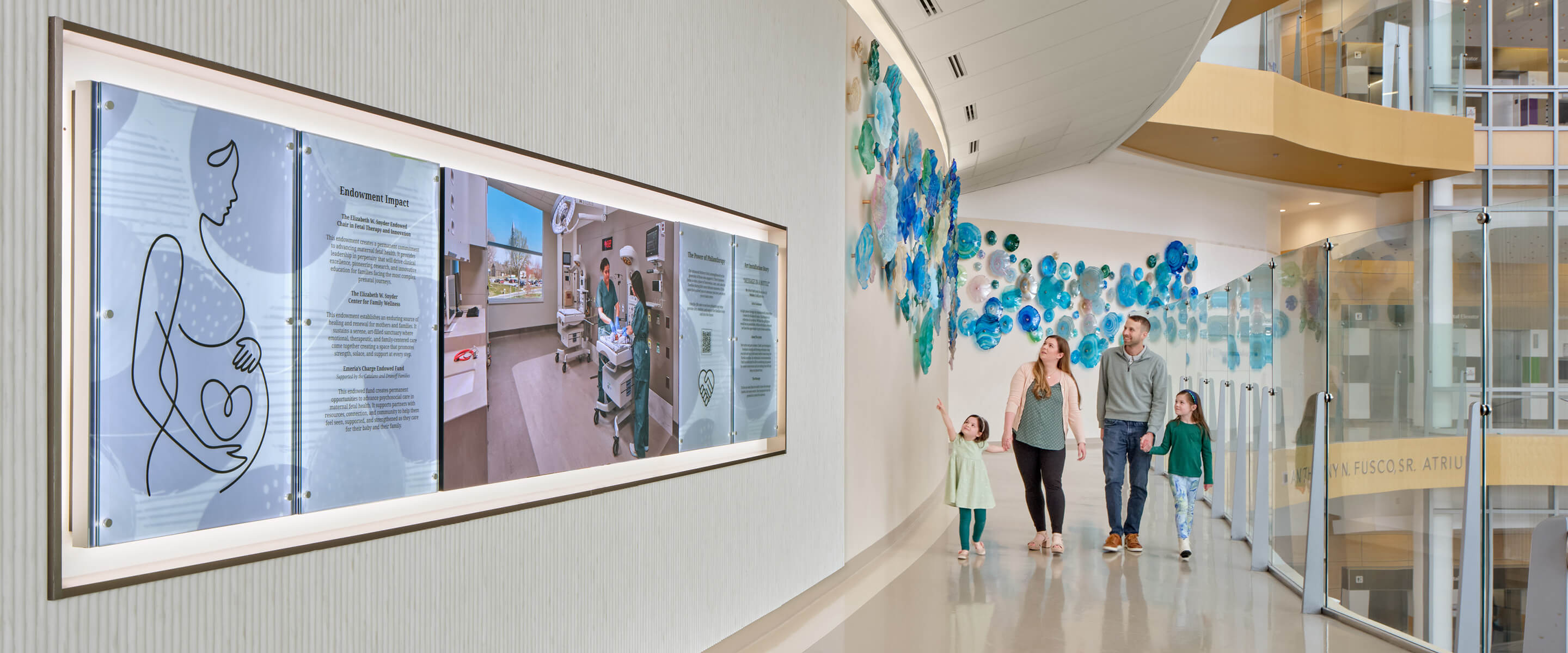 Family walking in a hospital corridor, observing art displays and information boards about maternal health initiatives.
