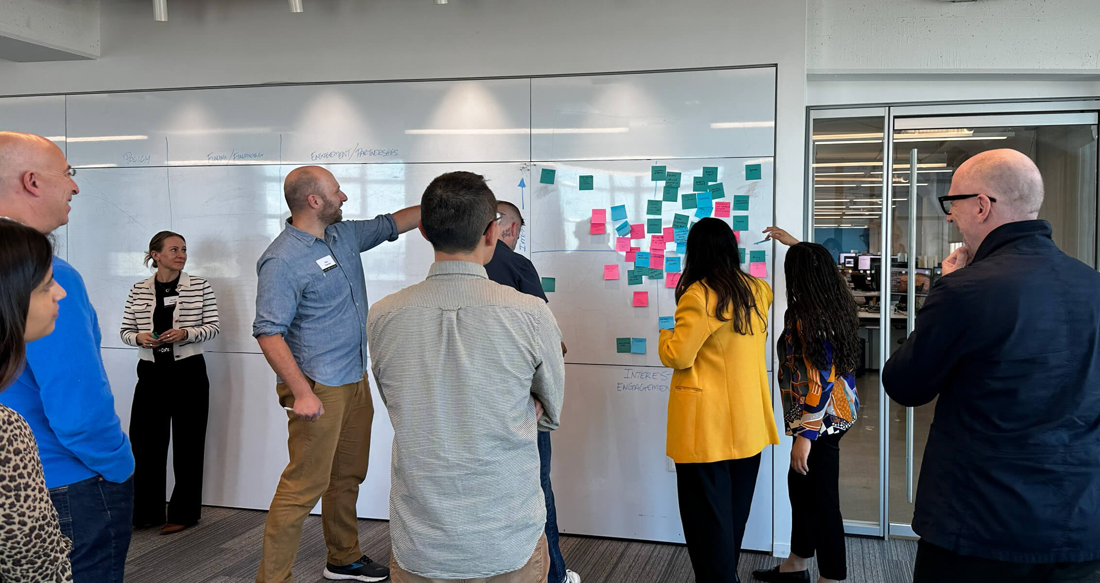 Group of people engaging in a brainstorming session by a whiteboard covered in colorful sticky notes in an office setting.
