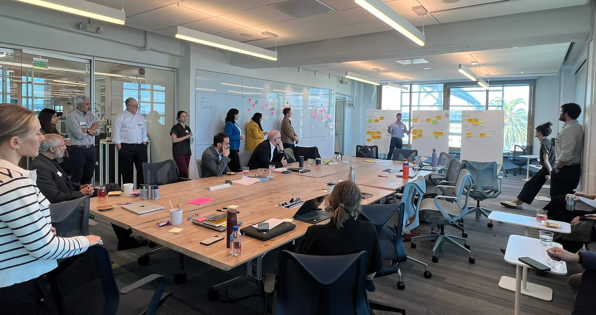 Group meeting in a bright conference room with participants engaged in discussion, surrounded by whiteboards and notes.