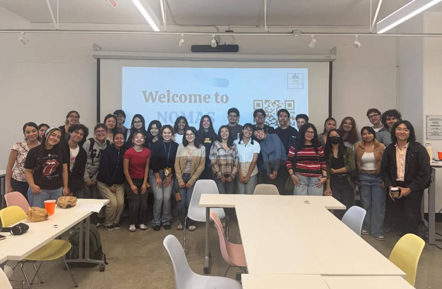 Group of students posing in a classroom with a presentation screen saying Welcome to NOMAS. Tables and chairs visible in the room.