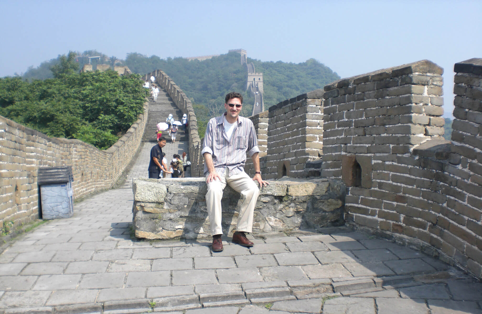 Basem sitting on a stone wall at the Great Wall of China, with visitors walking along the historic path and green hills in the background.