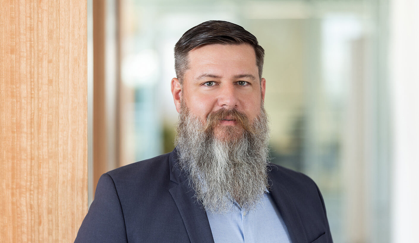 A bearded man in a suit stands near a wooden panel in an office setting, looking directly at the camera with a neutral expression.