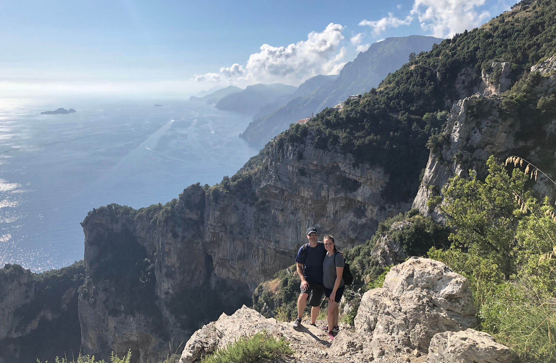 Lauren and her husband standing on a rocky cliff with a scenic view of the sea and mountains along the coastline, in bright daylight.