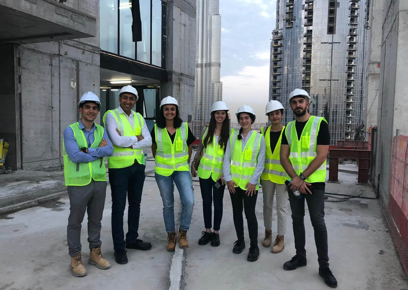 Razan and fellow project team members wearing safety vests and hard hats pose on a building site, with cranes and unfinished buildings in the background.
