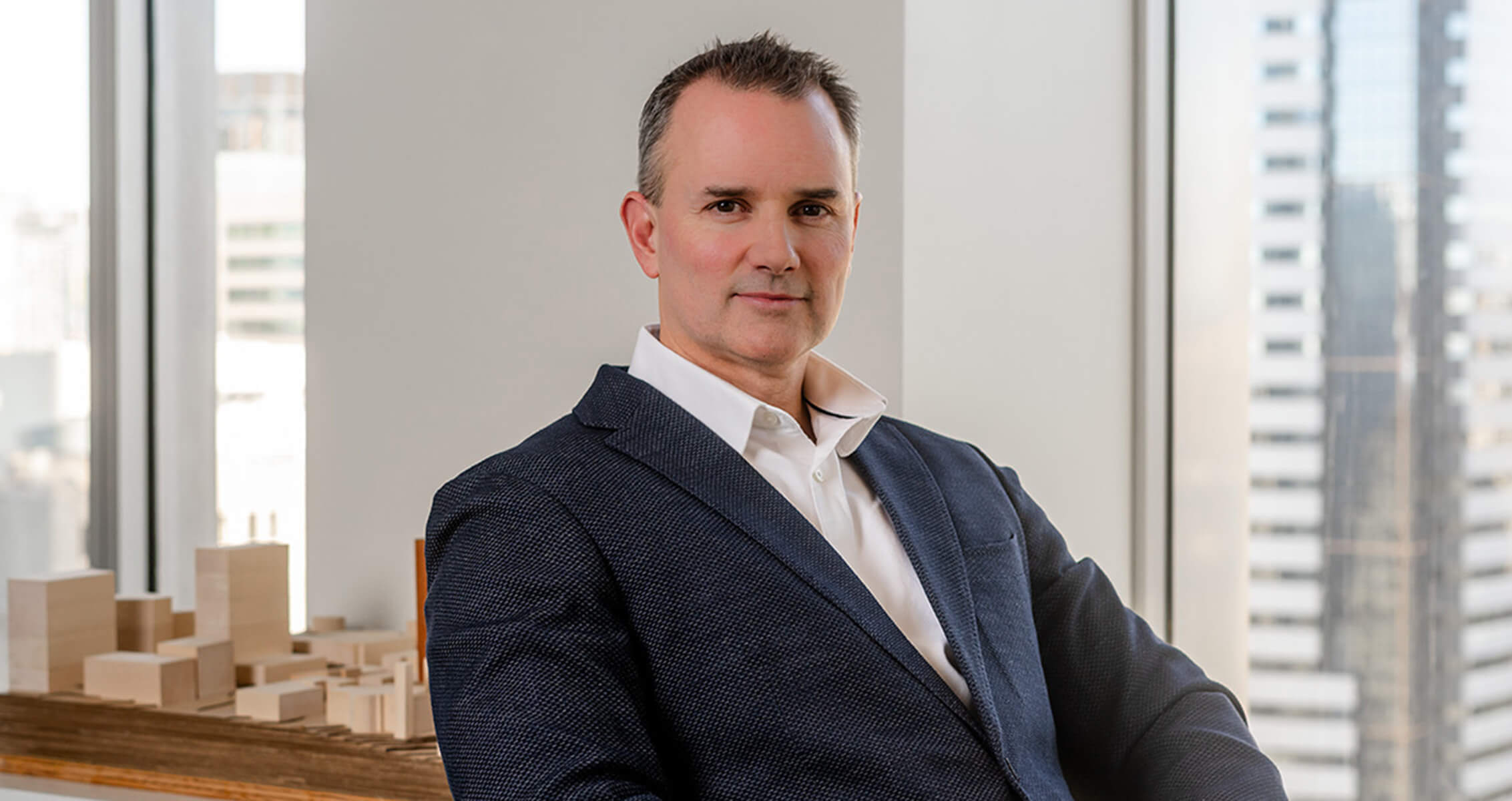 Portrait of a man in a dark blazer sitting in an office with a cityscape view, featuring architectural models in the background.