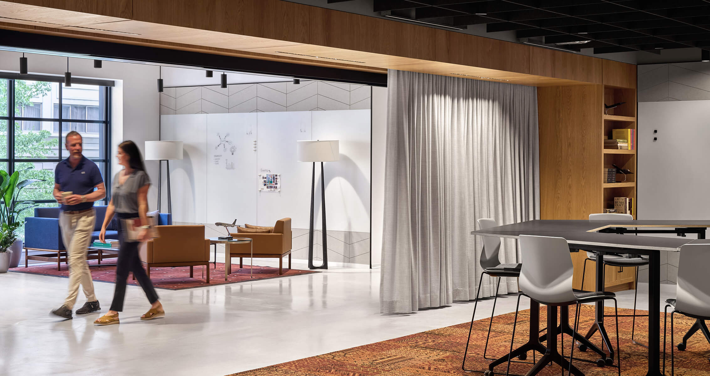 Two people walk through a contemporary office space featuring seating areas, a large window, and a discussion area with a table and chairs.