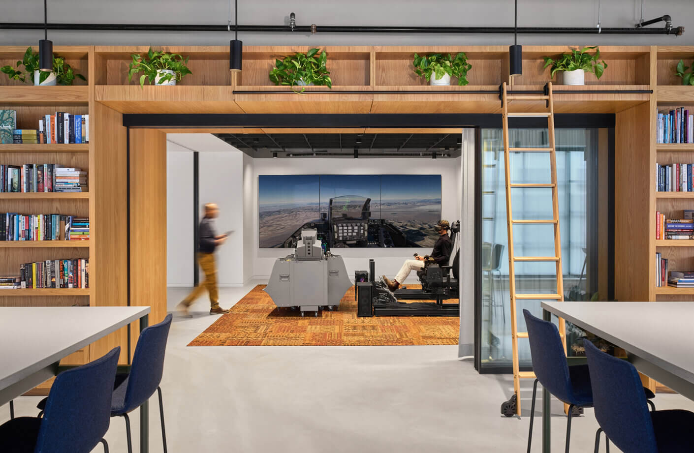 A workspace featuring a virtual reality flight simulator, bookshelves, a ladder, and a casually dressed person walking by.