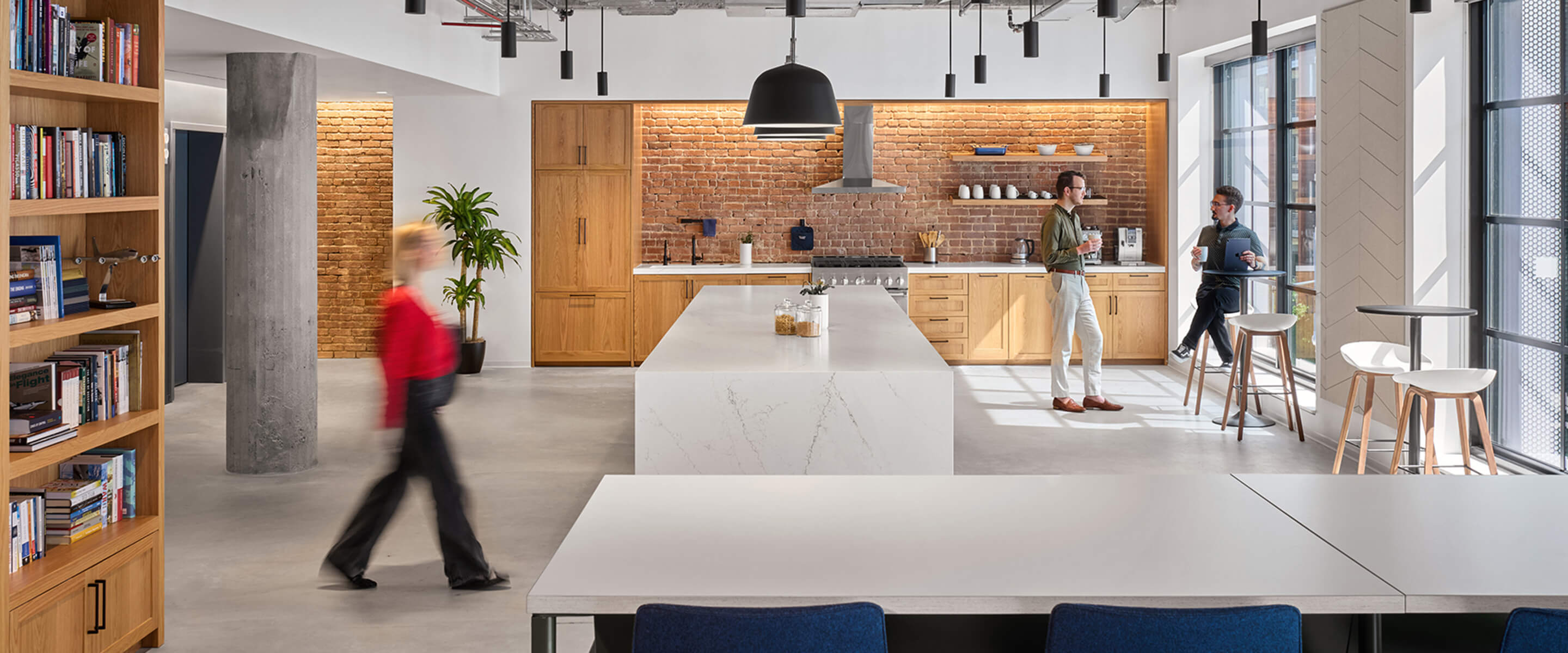 Bright open kitchen area with wood cabinetry, large island, and contemporary seating. Two people engaged in conversation, bookshelves in view.