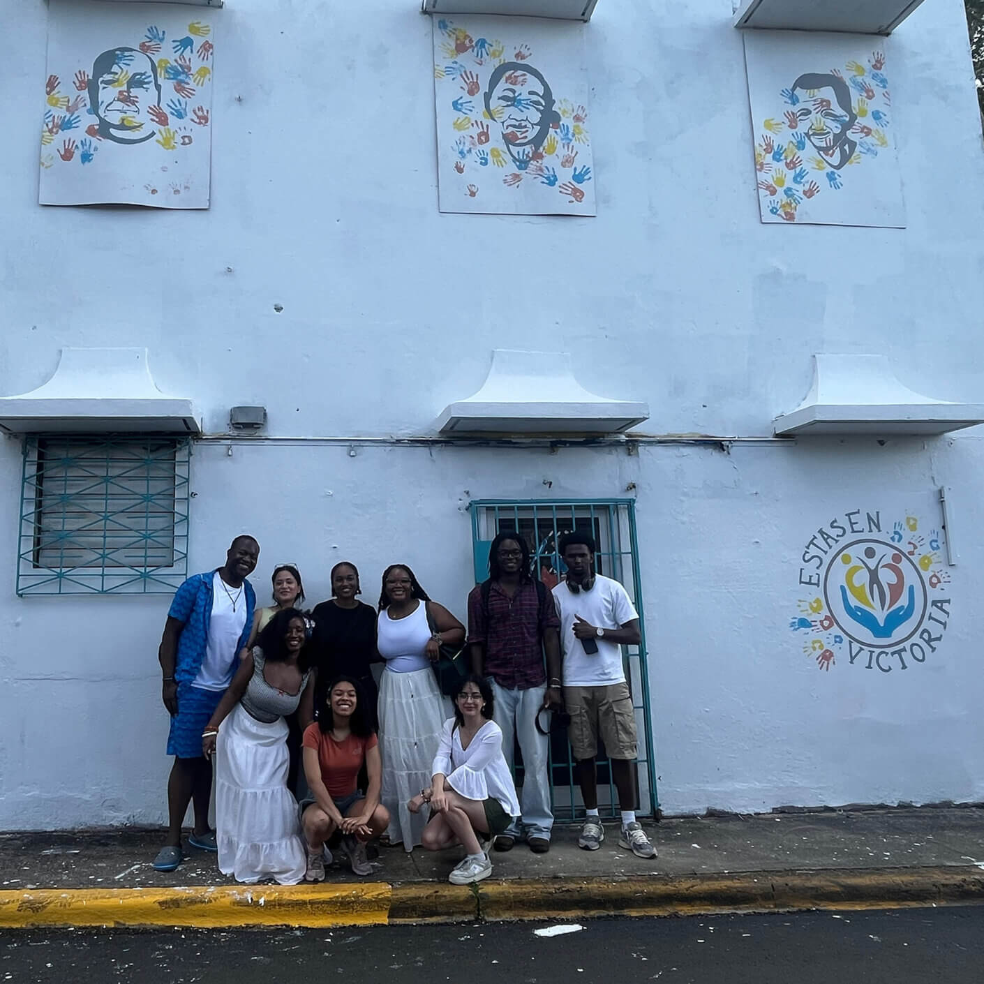 Group of diverse individuals posing in front of a white building with handprint art and the words Estás en Victoria.