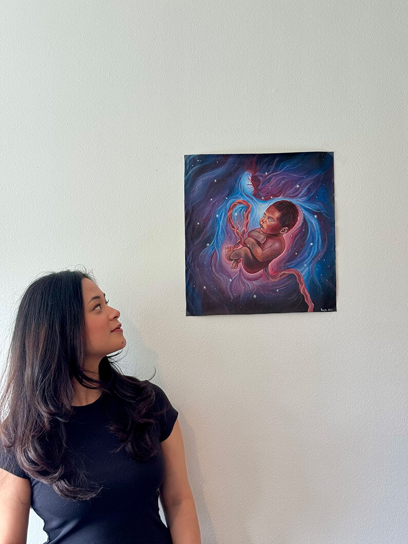 A woman in a black shirt looks at an artwork depicting a baby surrounded by cosmic elements on a light wall.