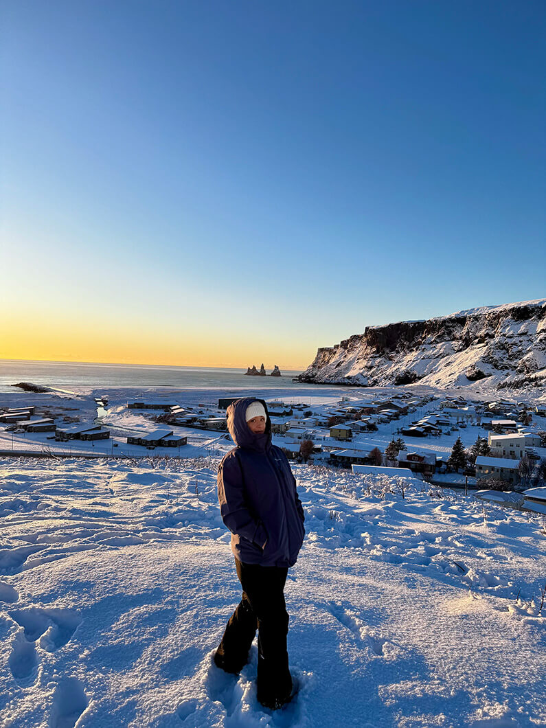 Person standing in snow-covered landscape, with a coastal village and cliffs in the background during sunset. Clear blue sky overhead.
