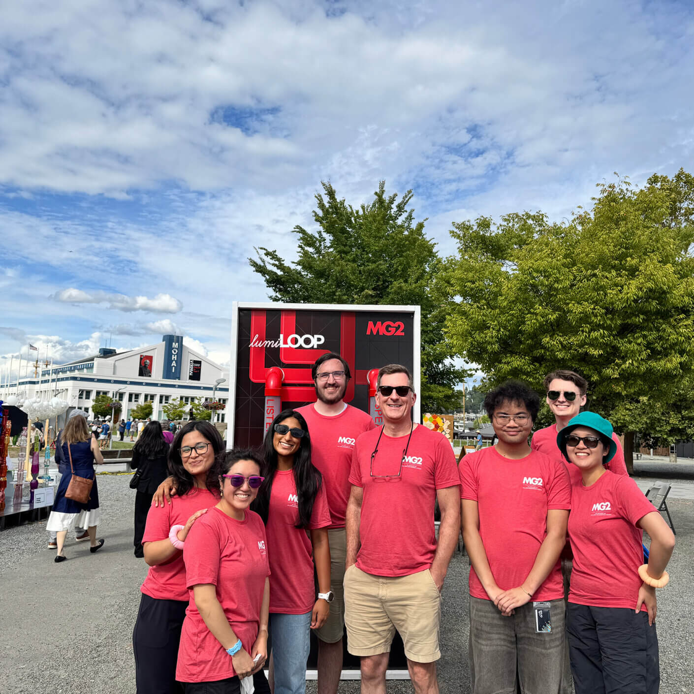 Group of people wearing red shirts standing in front of an MG2 sign at an outdoor event. Trees and buildings visible in the background.