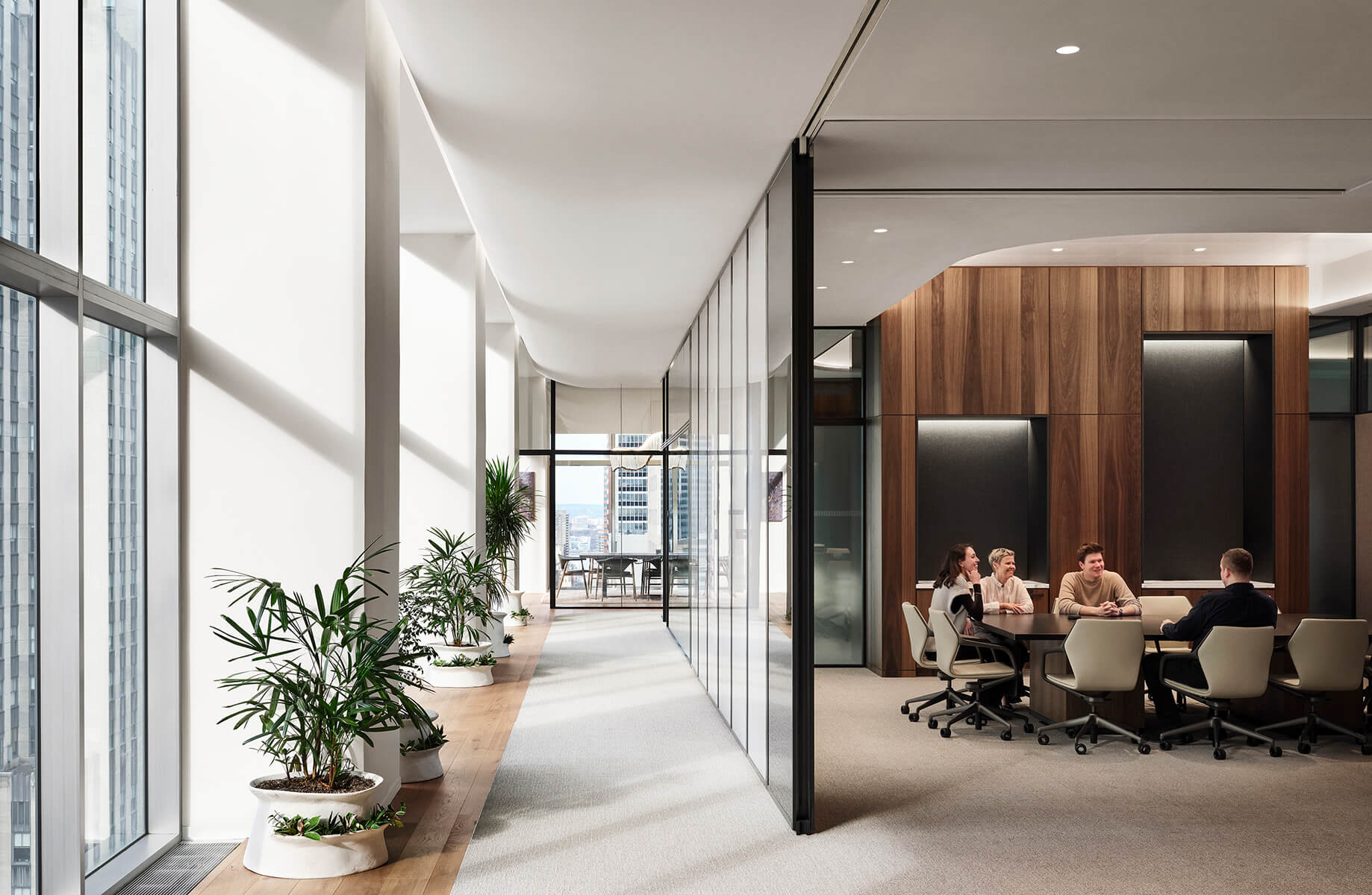 Office interior featuring glass walls, natural light, and plants. A meeting room with five people gathered around a table.