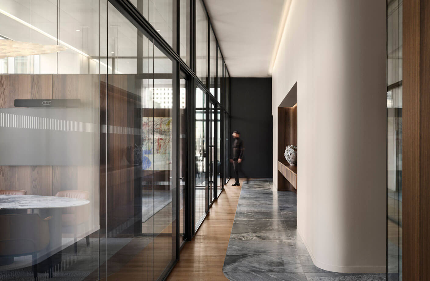 A sleek corridor featuring glass walls, wooden accents, and a marble floor, with a person walking past a decorative shelf.