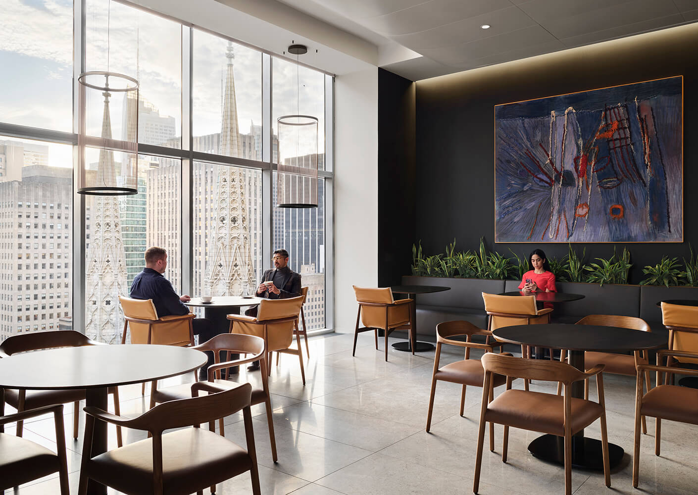 Interior seating area with large windows showcasing city views. Two men converse at a table, while a woman uses her phone in the corner.