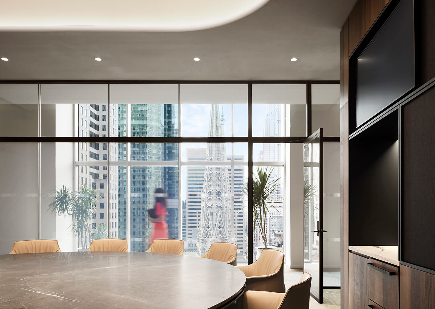 A conference room with a circular table and beige chairs, featuring large windows overlooking a city skyline with skyscrapers.