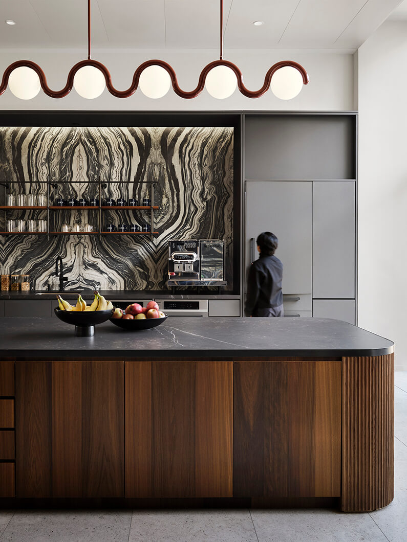 Stylish kitchen featuring a dark stone island, striped backdrop, and pendant lights, with a person preparing food and a fruit bowl on the counter.