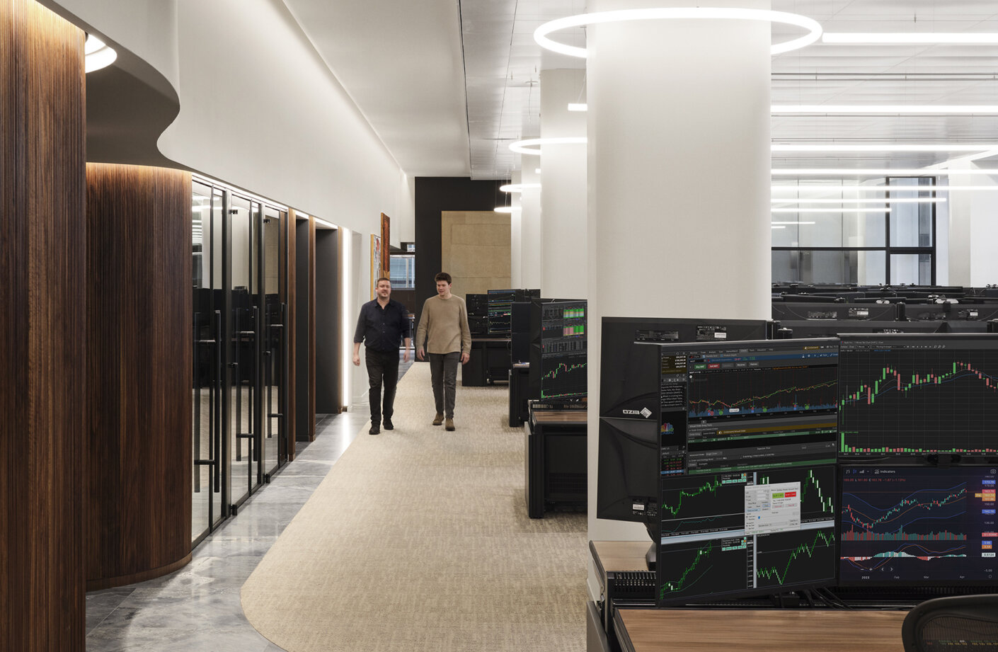 Two professionals walk through a bright office space featuring multiple computer desks with financial market displays and a modern interior design.
