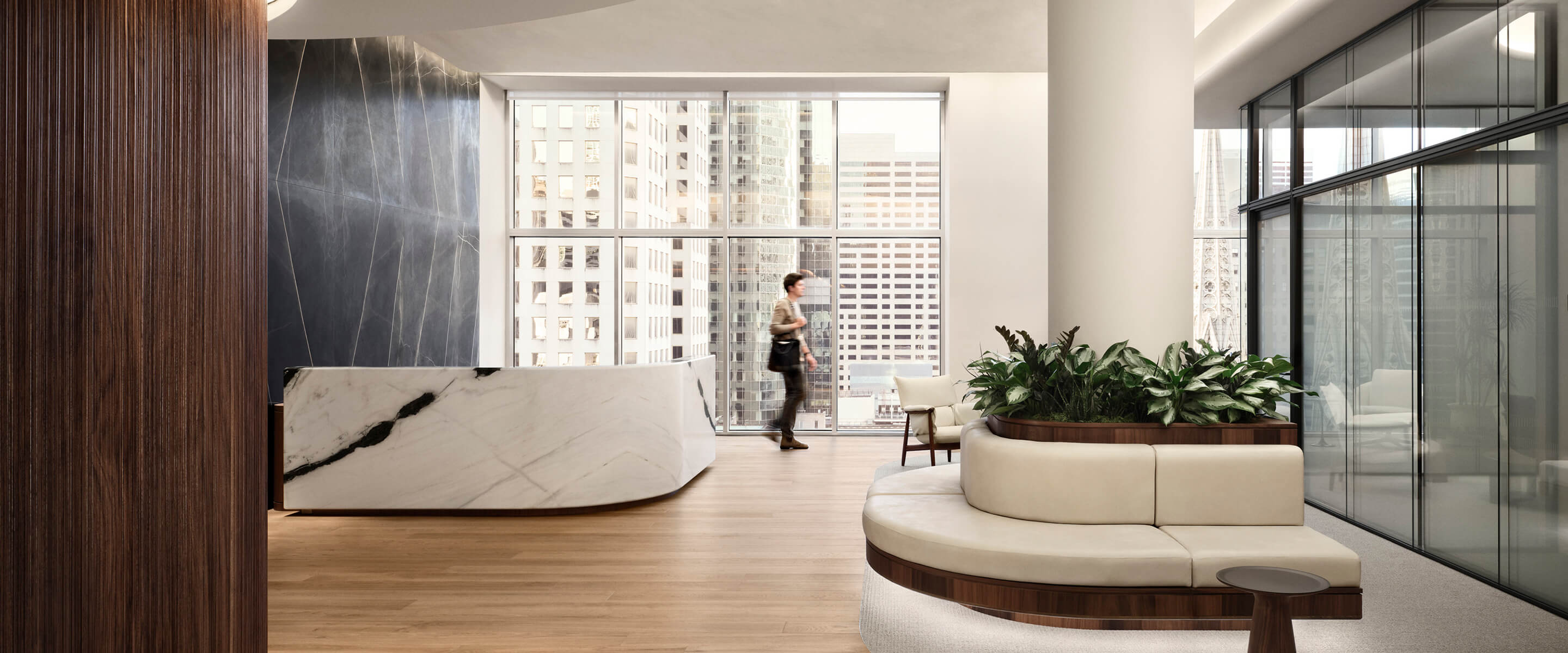 Office lobby featuring a marble reception desk, large windows with city views, and a lounge area with a circular sofa and plants.