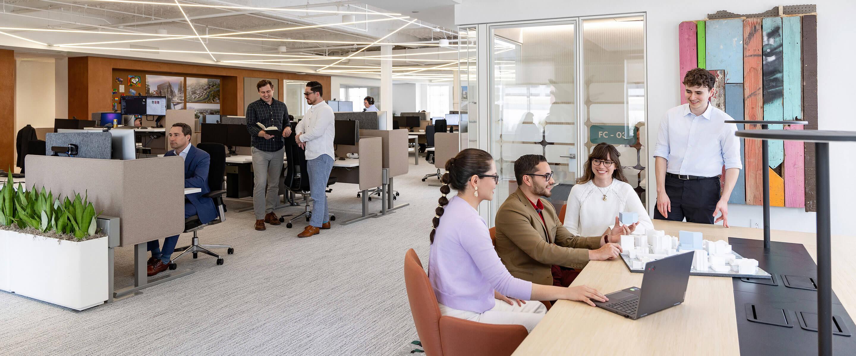 Group of professionals collaborating in an office space, discussing a project and reviewing a 3D model on a table, with desks in the background.
