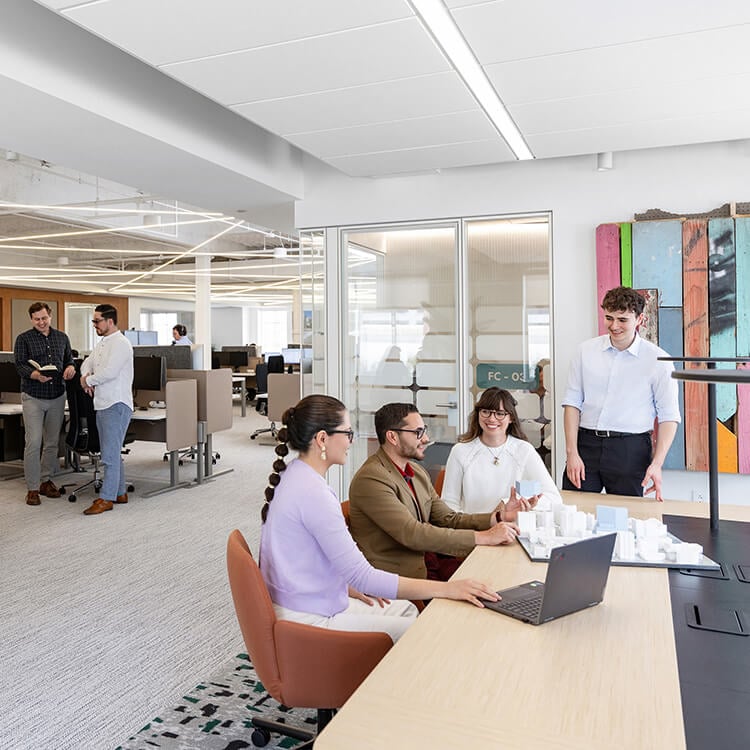 Group discussing architectural models in an office environment, with a mix of collaborative work and casual interactions among colleagues.