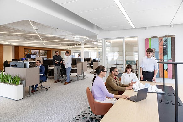 Office setting with employees collaborating at a desk, engaging in discussions, and working at computer stations in a spacious workspace.