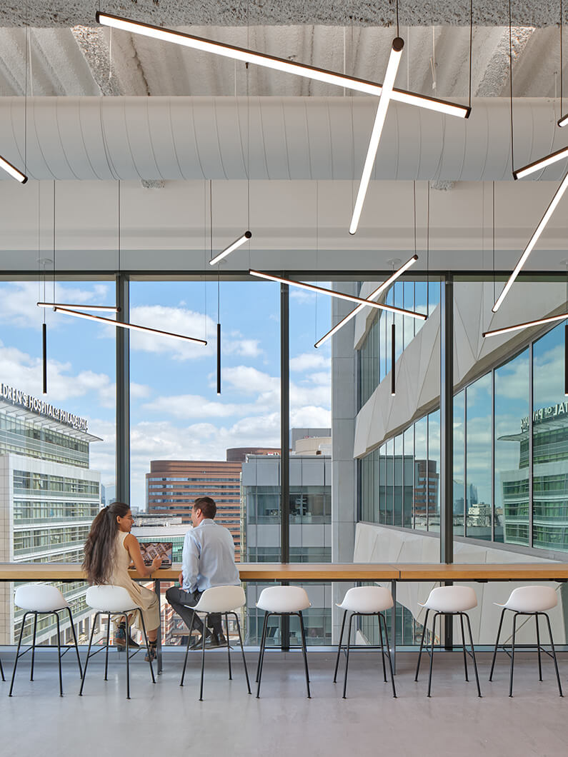 Two people sit at a long wooden table by large windows, discussing work in a bright office space with unique pendant lights overhead.