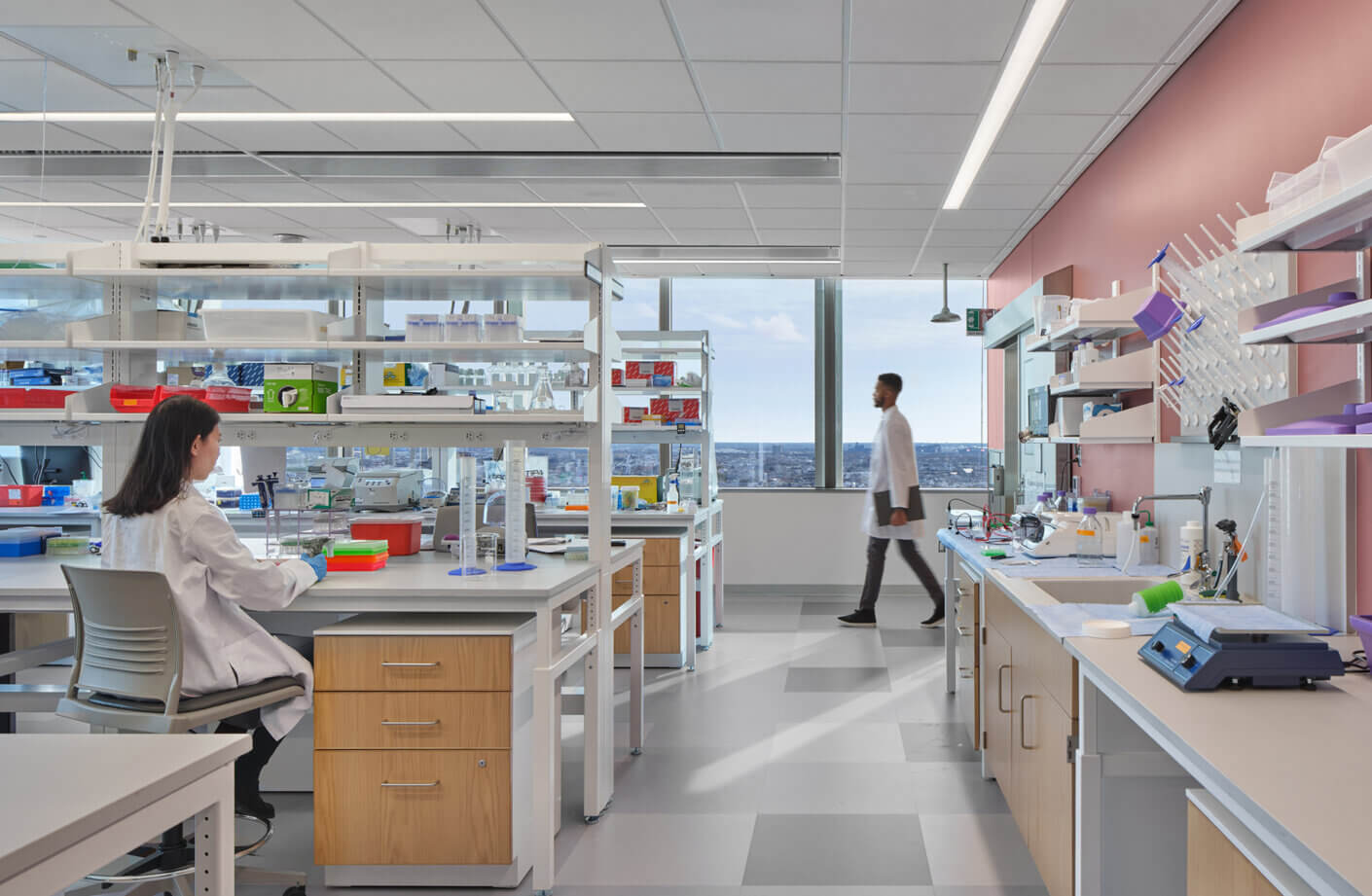 Laboratory space with two scientists, one seated and working at a lab bench, and another walking between workstations with equipment and supplies.