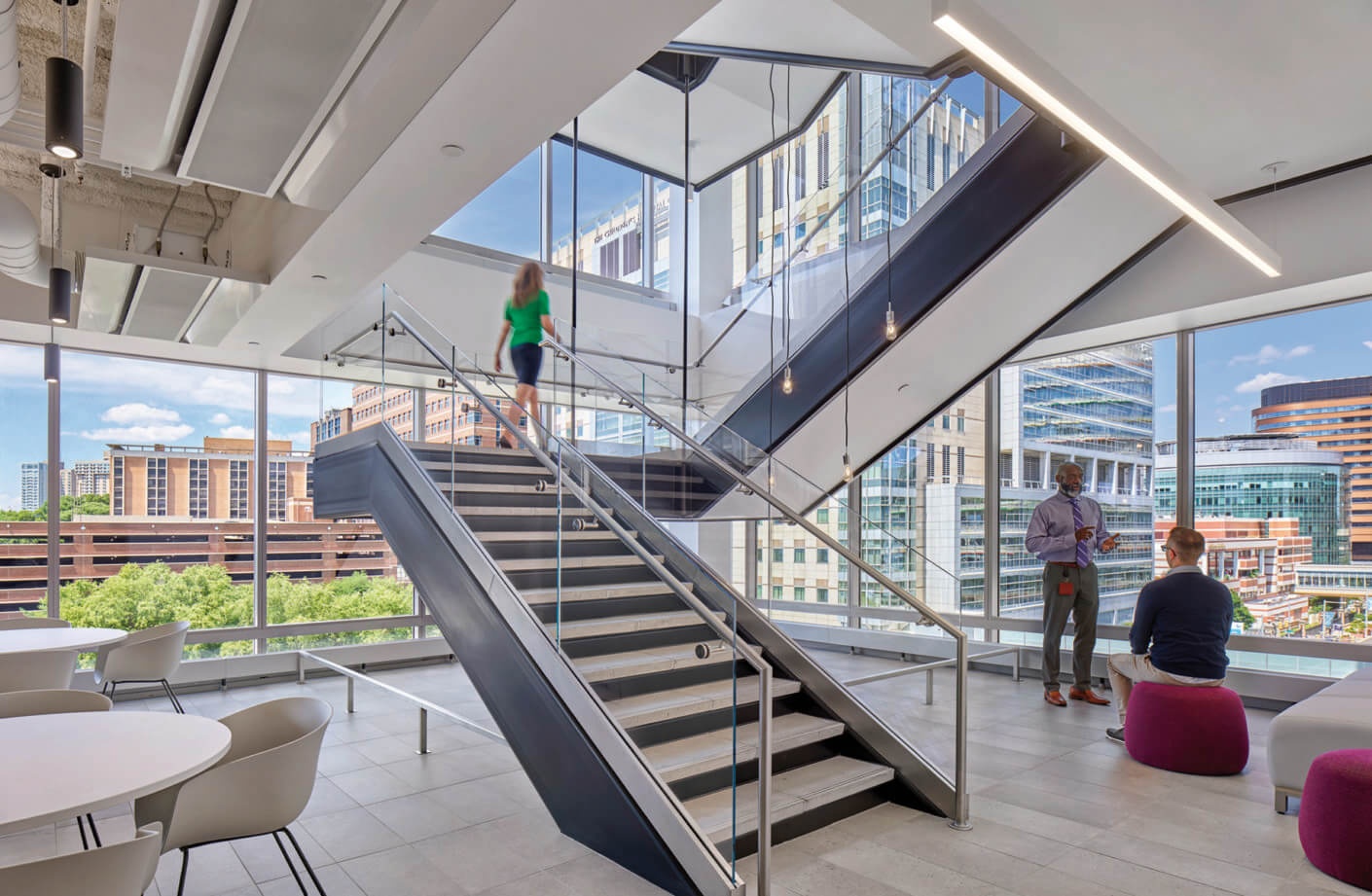 Interior view of a staircase with glass railings, showing people in conversation and large windows overlooking a cityscape and greenery.